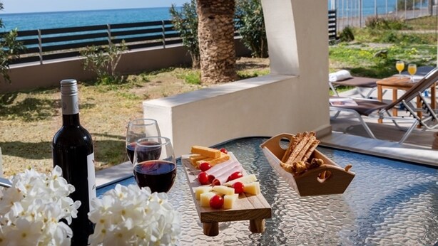 A table with a bottle of wine, glasses, cheese, and crackers, set on a balcony overlooking a beach and the sea. - Atalanti Seaside Junior Suites (Viannos)