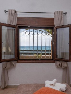 Ein Schlafzimmer mit großem Fenster und Meerblick, einem Bett mit orangefarbener Decke und Vorhängen an beiden Seiten.