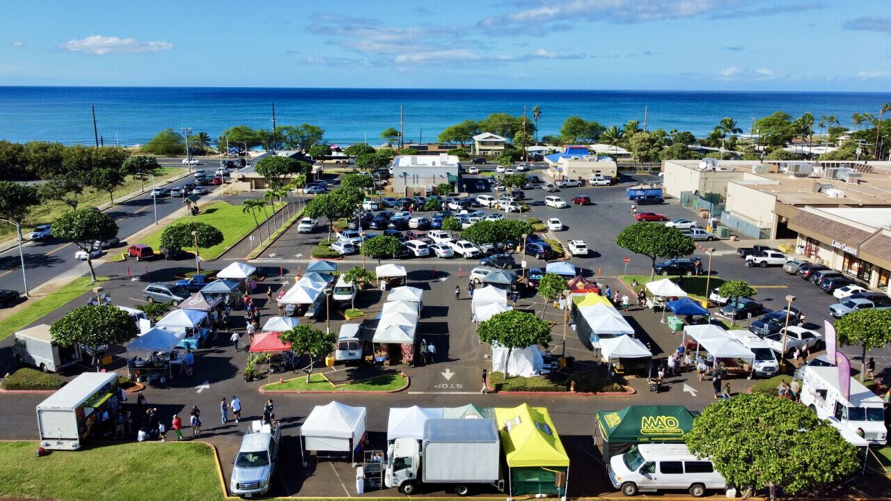 An aerial view of a parking lot with multiple tents and vehicles, likely a temporary event or market, with a clear view of the ocean in the background.