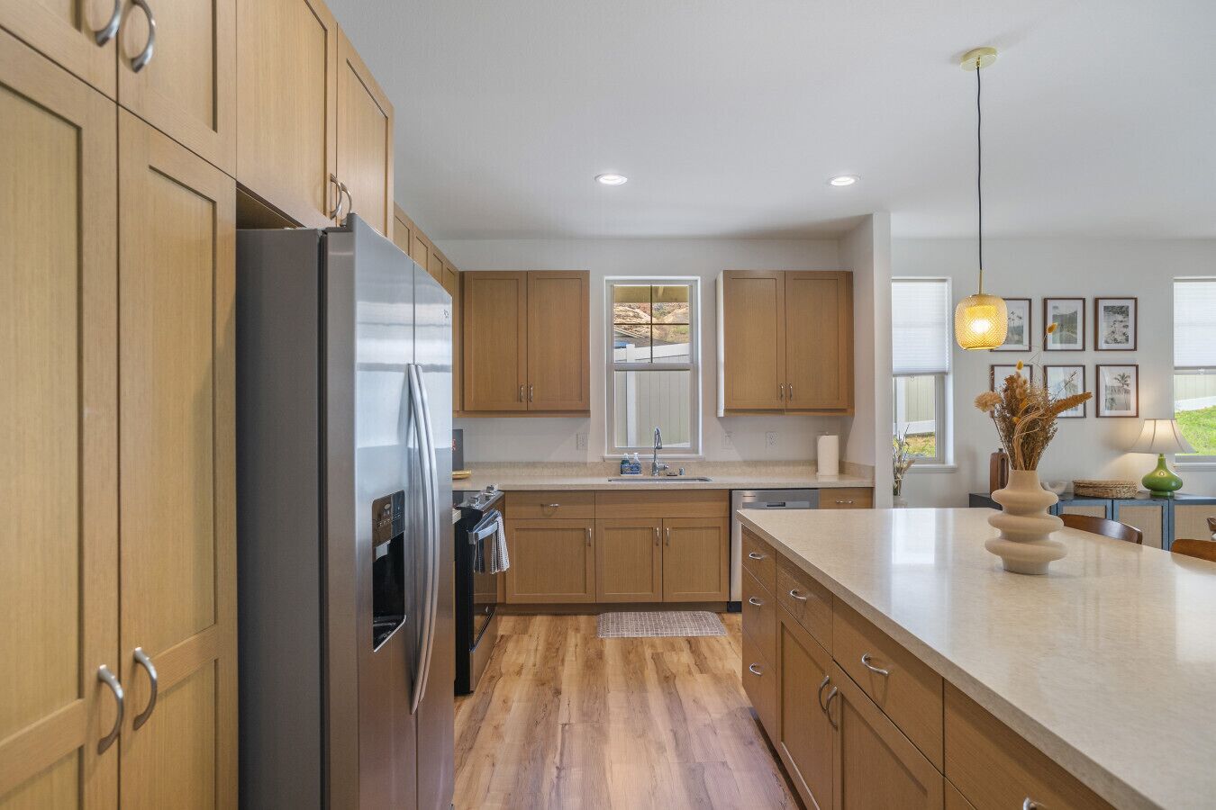 A modern kitchen with wooden cabinets, a stainless steel refrigerator, a large island, and a hanging pendant light.