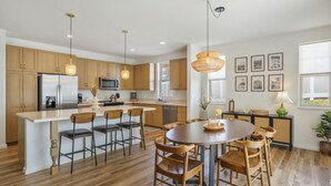 A modern kitchen with wooden cabinets, a central island with bar stools, a round dining table with chairs, pendant lighting, and framed artwork on the wall.