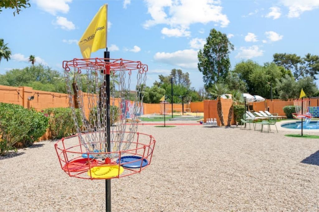 A disc golf basket with multiple colored discs, a flag, and a pool area in the background.