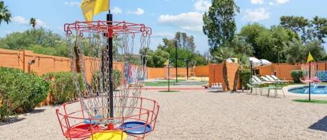 A disc golf basket with multiple colored discs, a flag, and a pool area in the background.