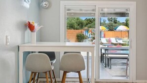Une cuisine moderne avec un plan de travail blanc, deux chaises et une porte vitrée donnant sur un patio aménagé avec du mobilier d’extérieur et des parasols.