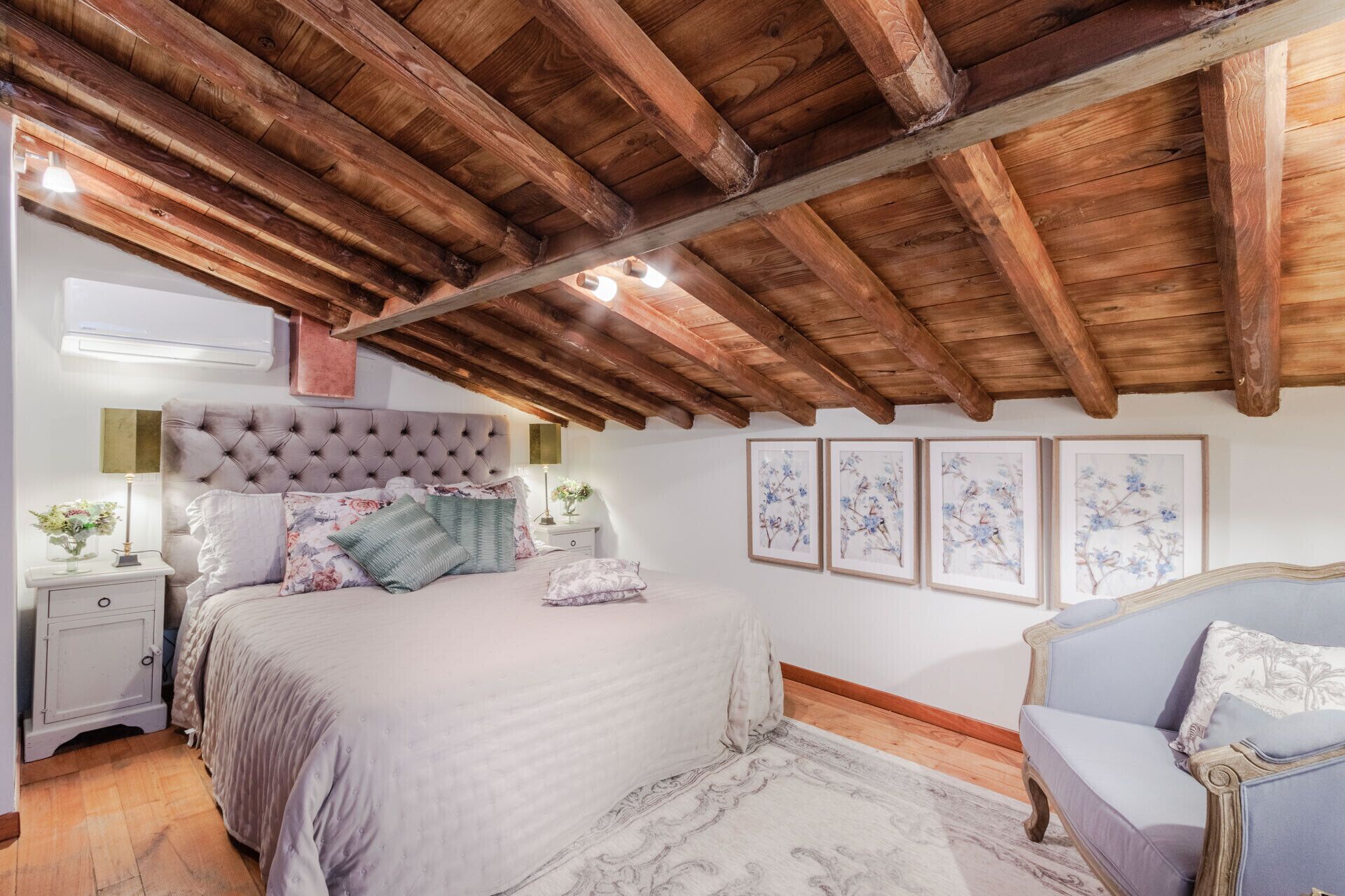 Ein Schlafzimmer mit Holzbalkendecke, einem großen Bett mit floralem Bettzeug, Nachttischen und gerahmten Kunstwerken an der Wand.