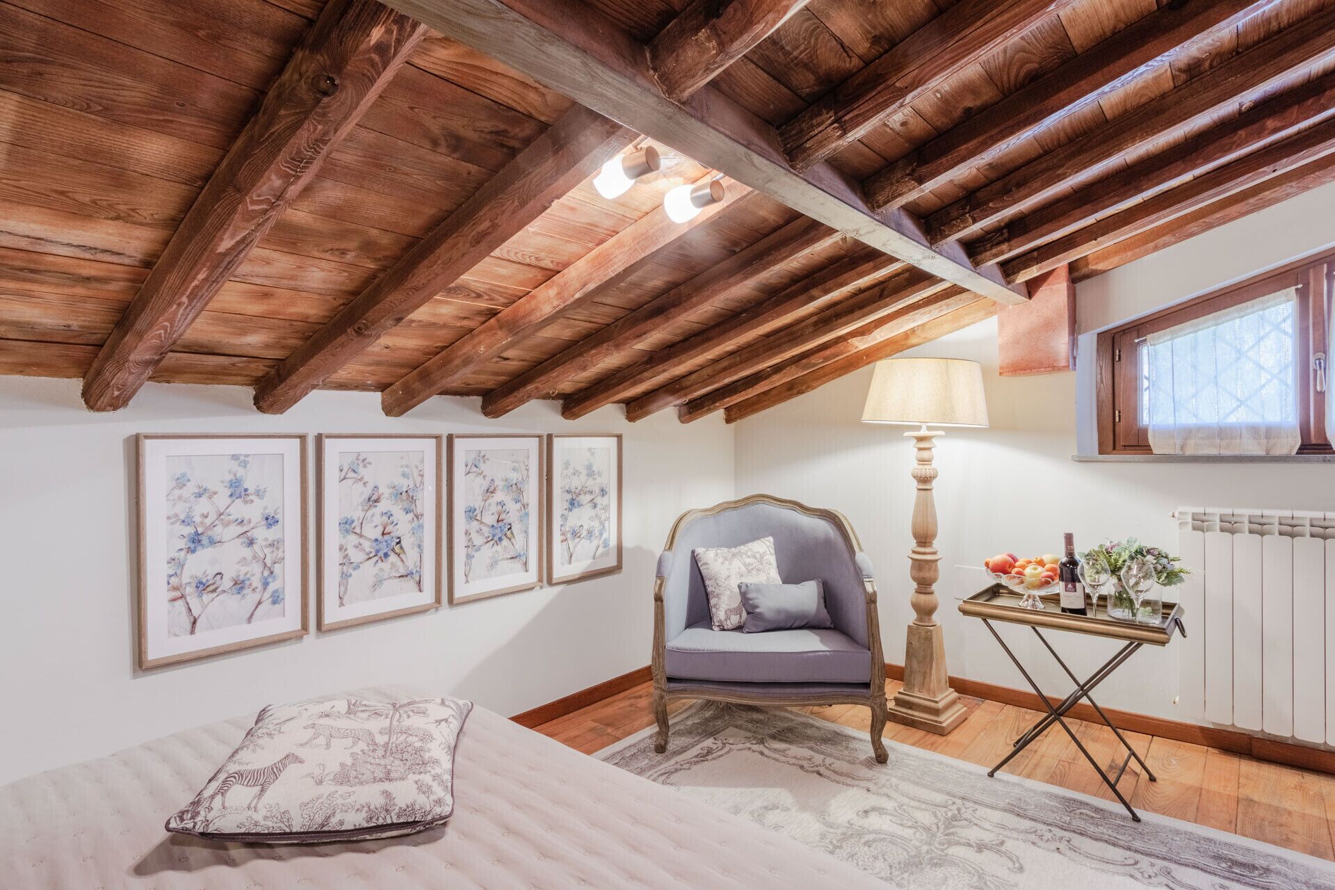 A cozy room with a wooden ceiling, a bed with a floral pillow, a chair, a table with a lamp, and framed floral artwork on the wall.
