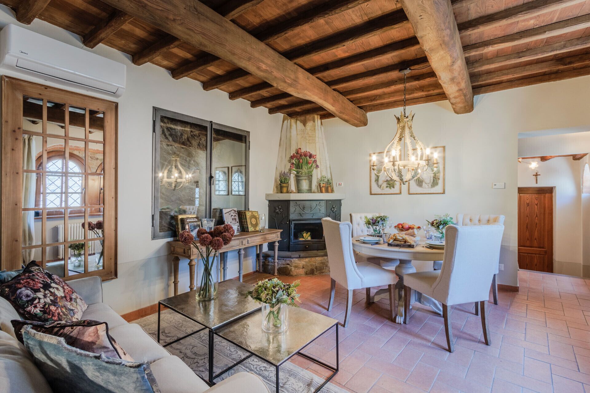 A cozy living room with a fireplace, wooden ceiling beams, and a dining area with a chandelier.