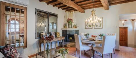 A cozy living room with a fireplace, wooden ceiling beams, and a dining area with a chandelier.