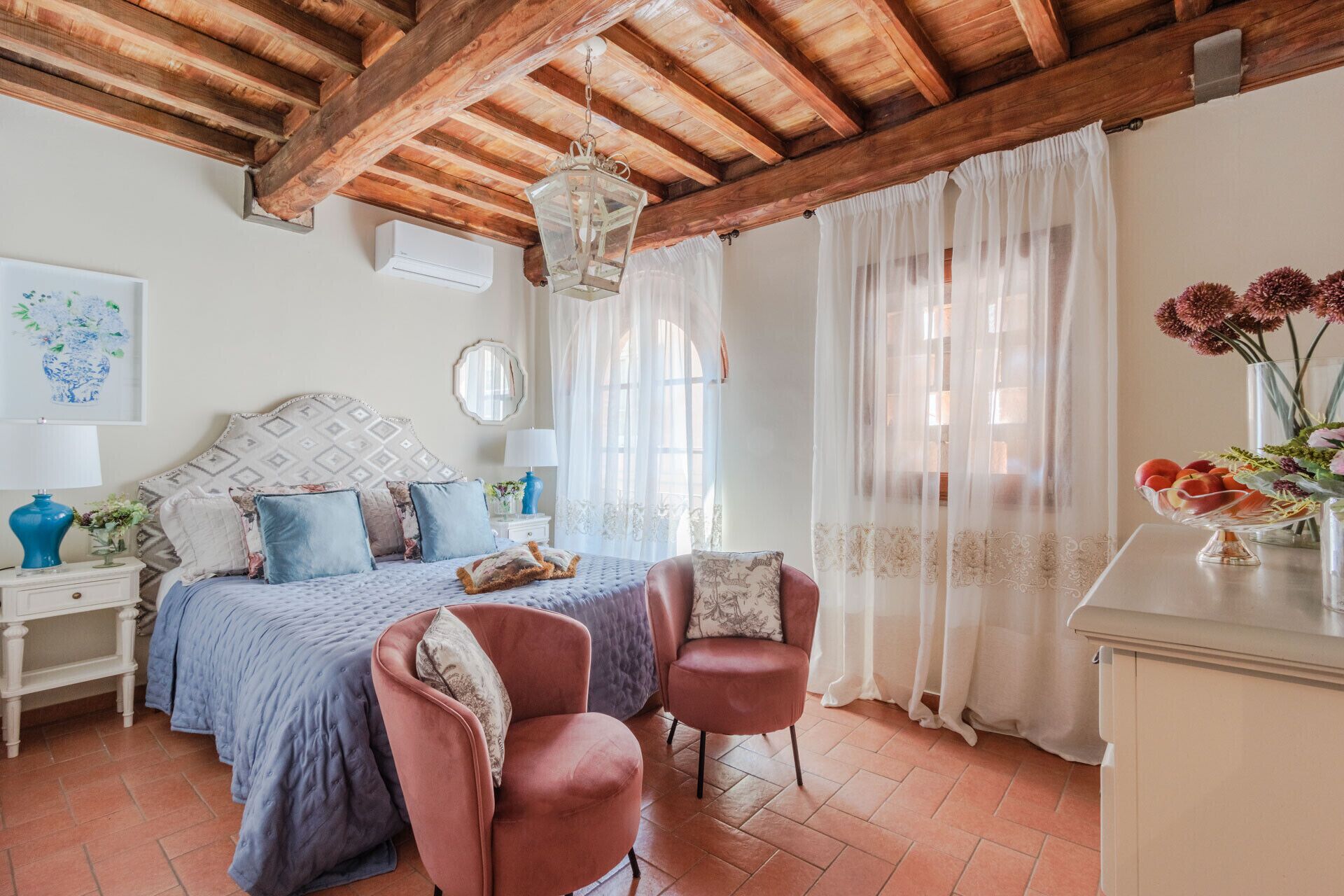 A bedroom with a bed, two chairs, a nightstand, a dresser, and a window with sheer curtains.