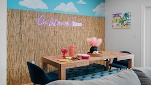 Um quarto com parede de madeira, um letreiro neon com a inscrição "On Island Town", uma mesa com cadeiras e um quadro na parede.
