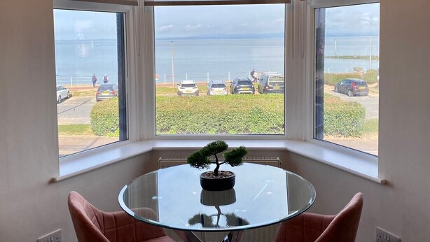 Quarto com mesa de vidro e duas cadeiras, uma árvore bonsai sobre a mesa, e vista para um estacionamento e mar através de amplas janelas.