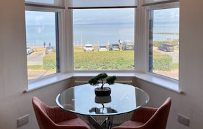 A room with a glass table and two chairs, a bonsai tree on the table, and a view of a parking area and sea through large windows. - Eden Retreats Serviced Apartments (Morecambe)