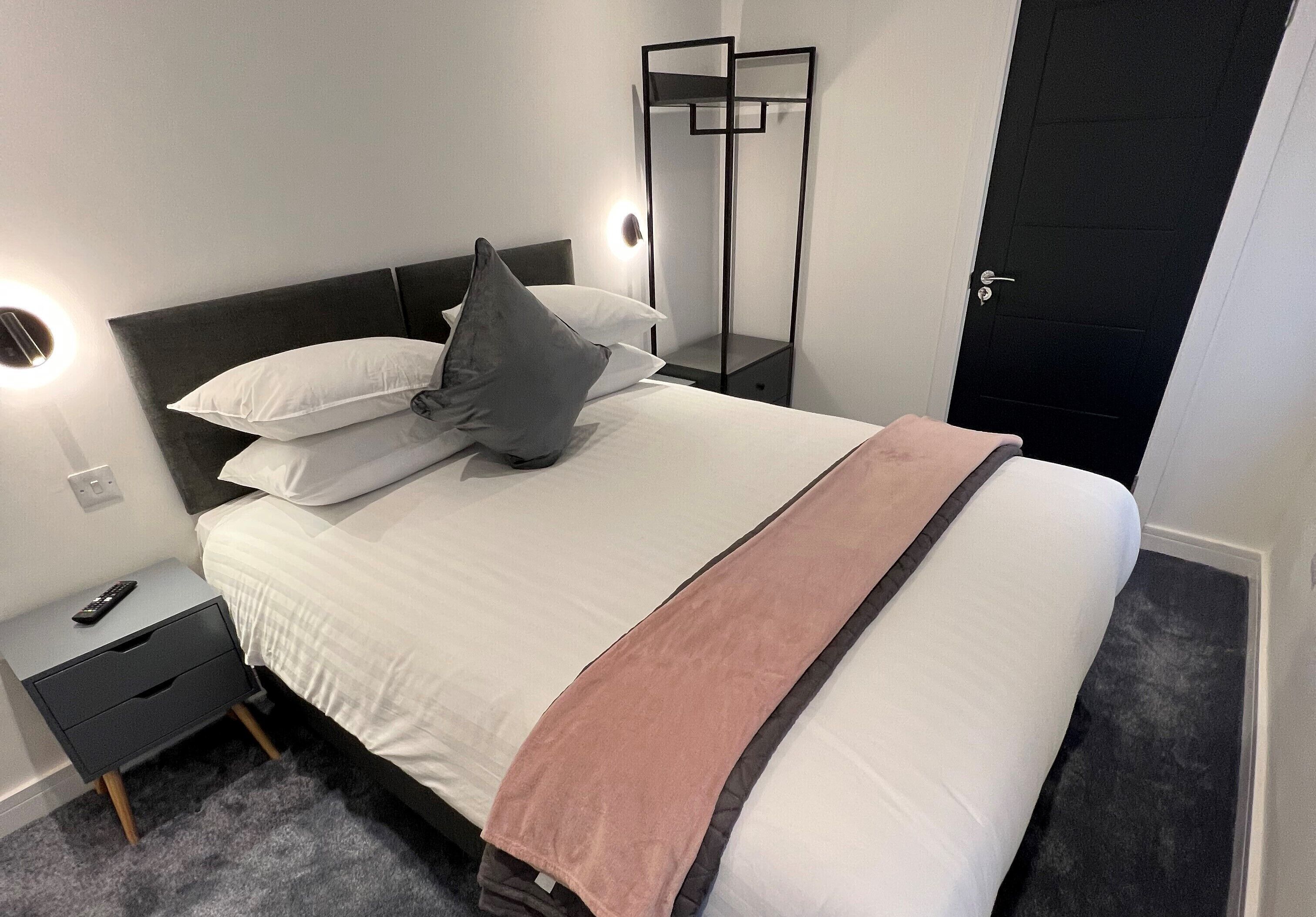 Ein ordentlich gemachtes Bett mit weißer Bettwäsche und einer rosafarbenen Tagesdecke, ein Nachttisch mit Fernbedienung und ein schwarzer Kleiderschrank mit Spiegel.