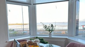 Quarto de canto com mesa de vidro e cadeira rosa, cesta de petiscos e vista para o mar através de grandes janelas.