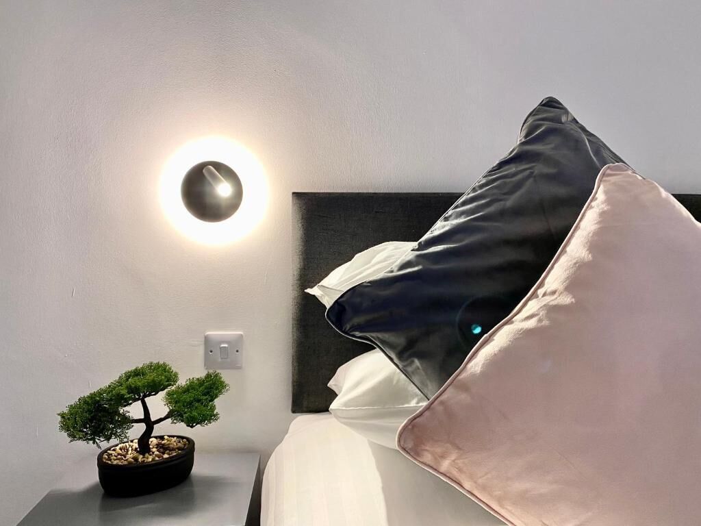 A neatly arranged hotel room with a bed, pillows, a bedside table with a bonsai tree, and a wall-mounted light fixture.