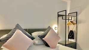 Cama bem arrumada com almofadas, mesa de cabeceira com abajur e espelho com bolsa pendurada.