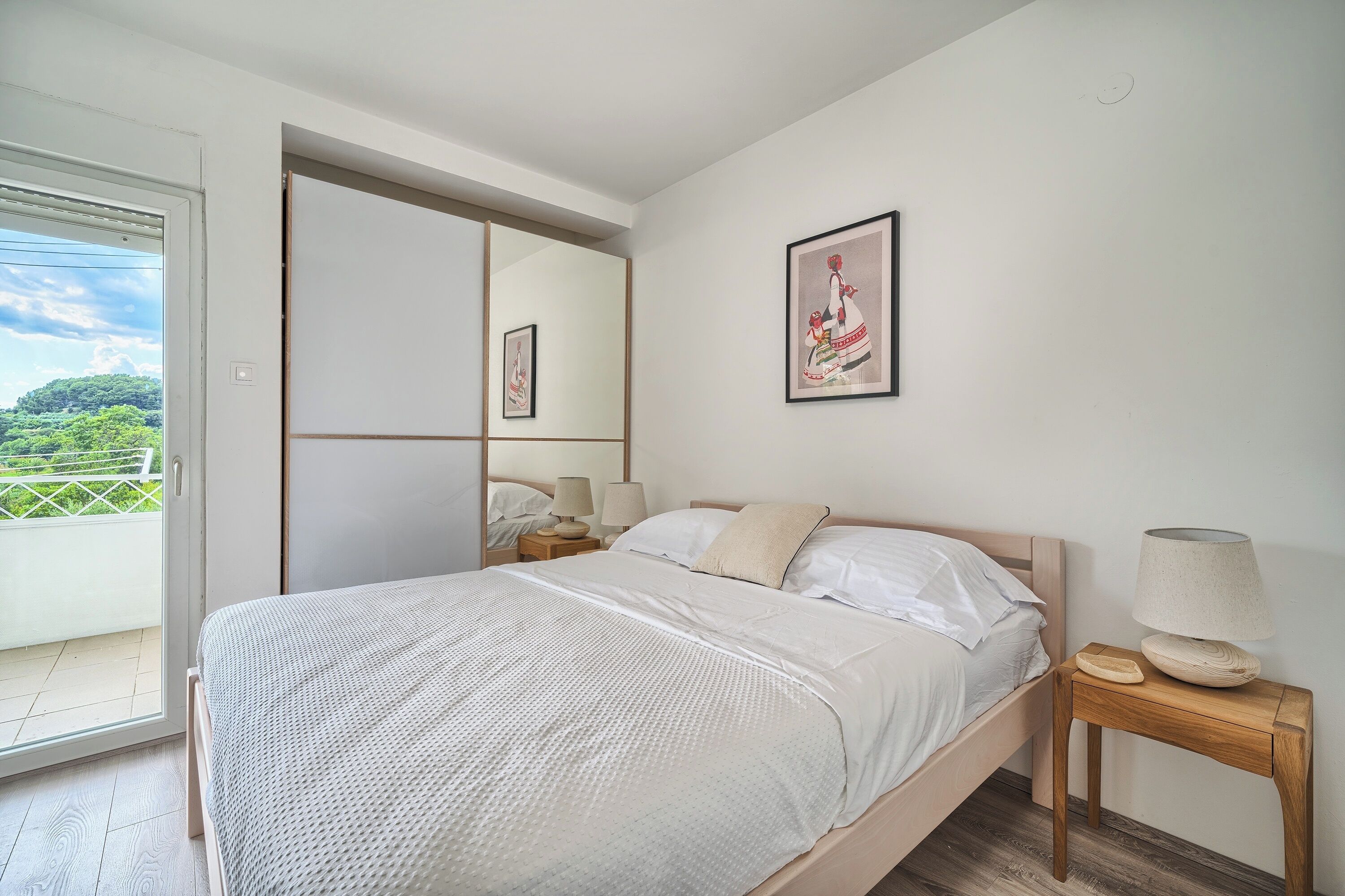 A bedroom with a bed, a nightstand, a lamp, a mirror, and a door leading to a balcony.