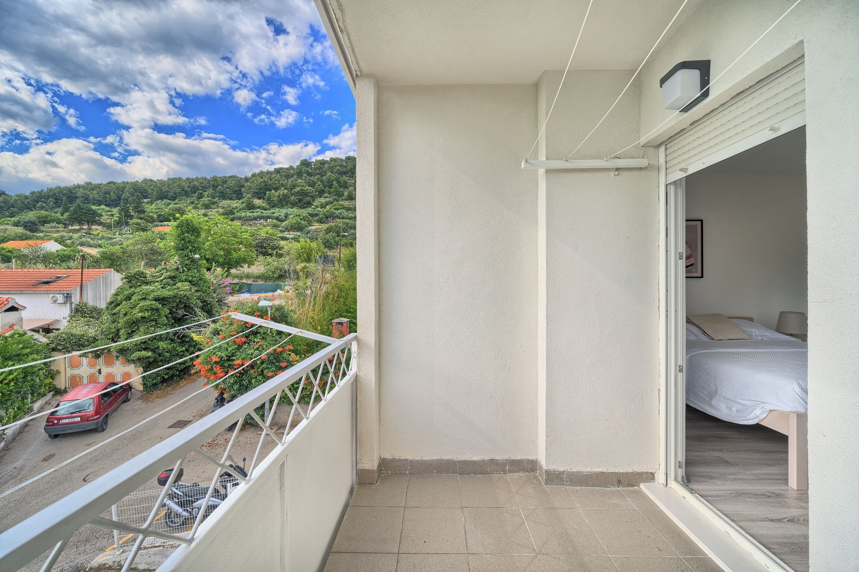 Un balcón con vista a una carretera y vegetación, un dormitorio con cama y mesita de noche, y una pared blanca con una lámpara.