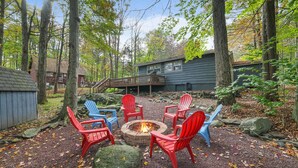 A fire pit surrounded by red and blue Adirondack chairs in a wooded area with a house in the background.