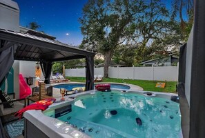 Relax in the hot tub surrounded by a lush lawn and a tree, with lounge chairs and a table nearby.