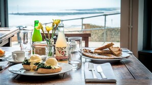 En frukostuppläggning med ägg Benedict, rostat bröd och en flaska äppeljuice på ett bord med utsikt över havet.