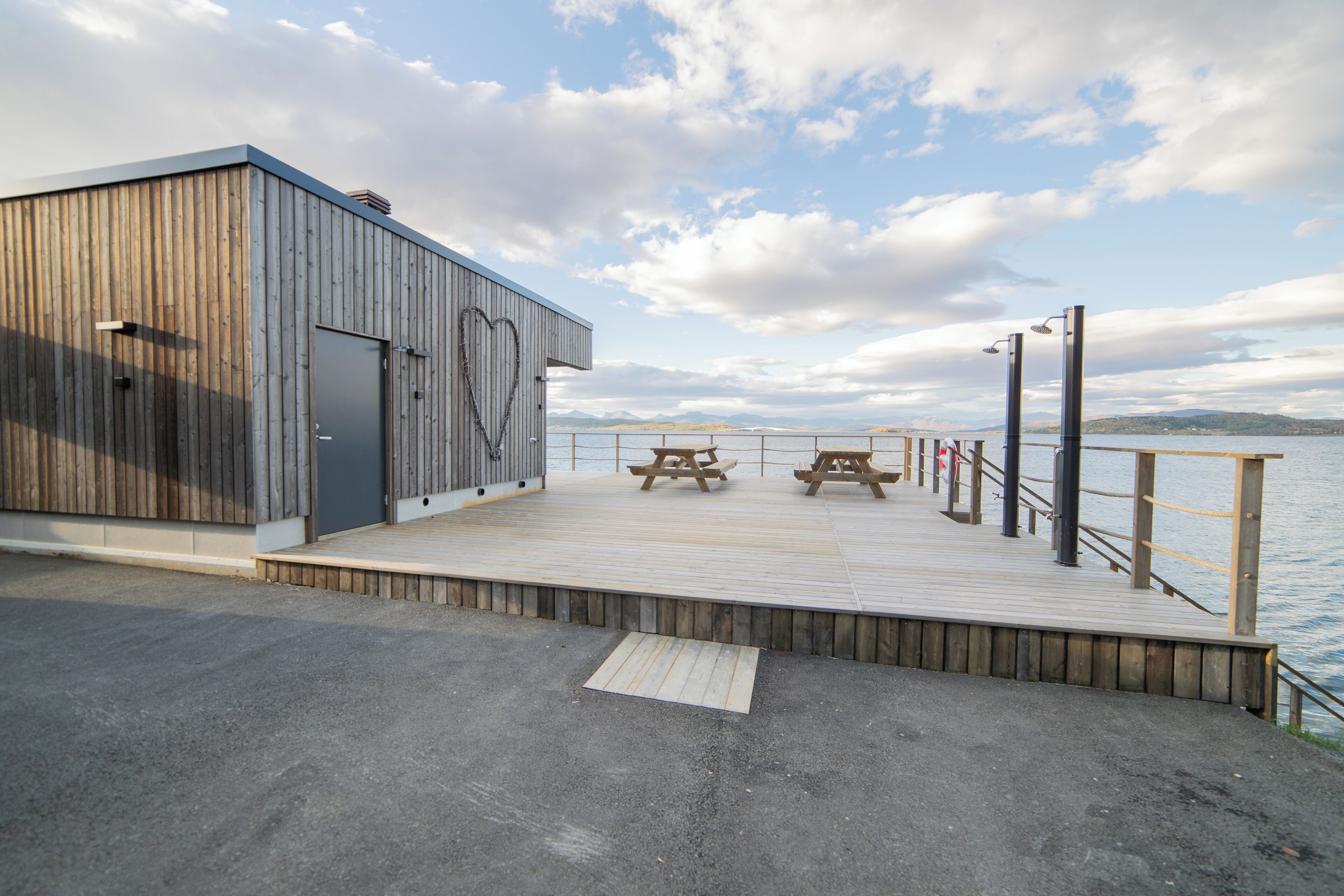 Una terraza de madera con mesas de picnic con vista a un cuerpo de agua, un edificio de metal con un ventanal en forma de corazón y un cielo despejado.