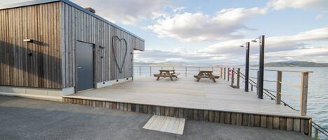Una terraza de madera con mesas de picnic con vista a un cuerpo de agua, un edificio de metal con un ventanal en forma de corazón y un cielo despejado.