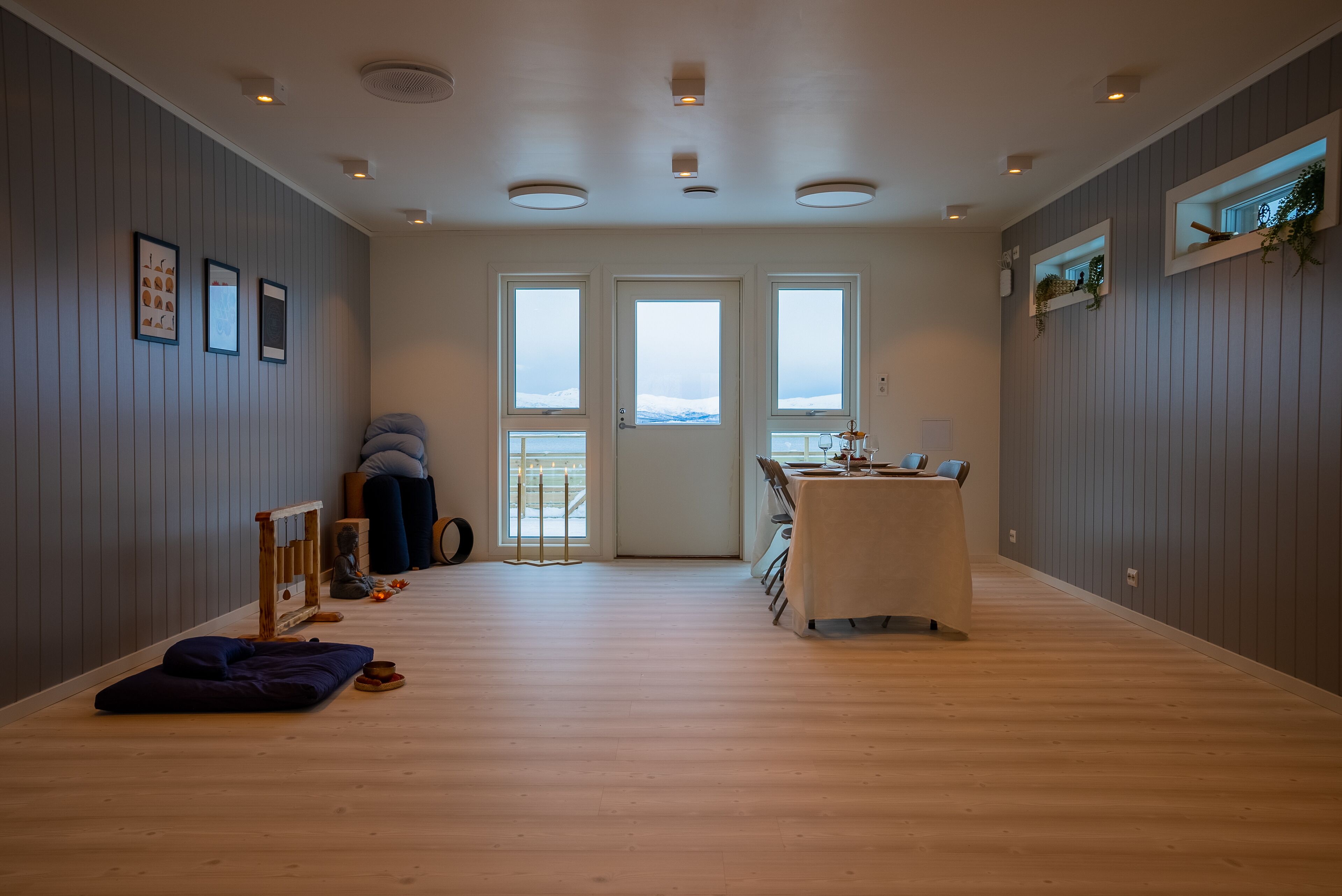 Una habitación amplia con suelo de madera, una mesa de comedor con sillas, una pequeña zona de estar con cojines y vistas a través de la puerta.