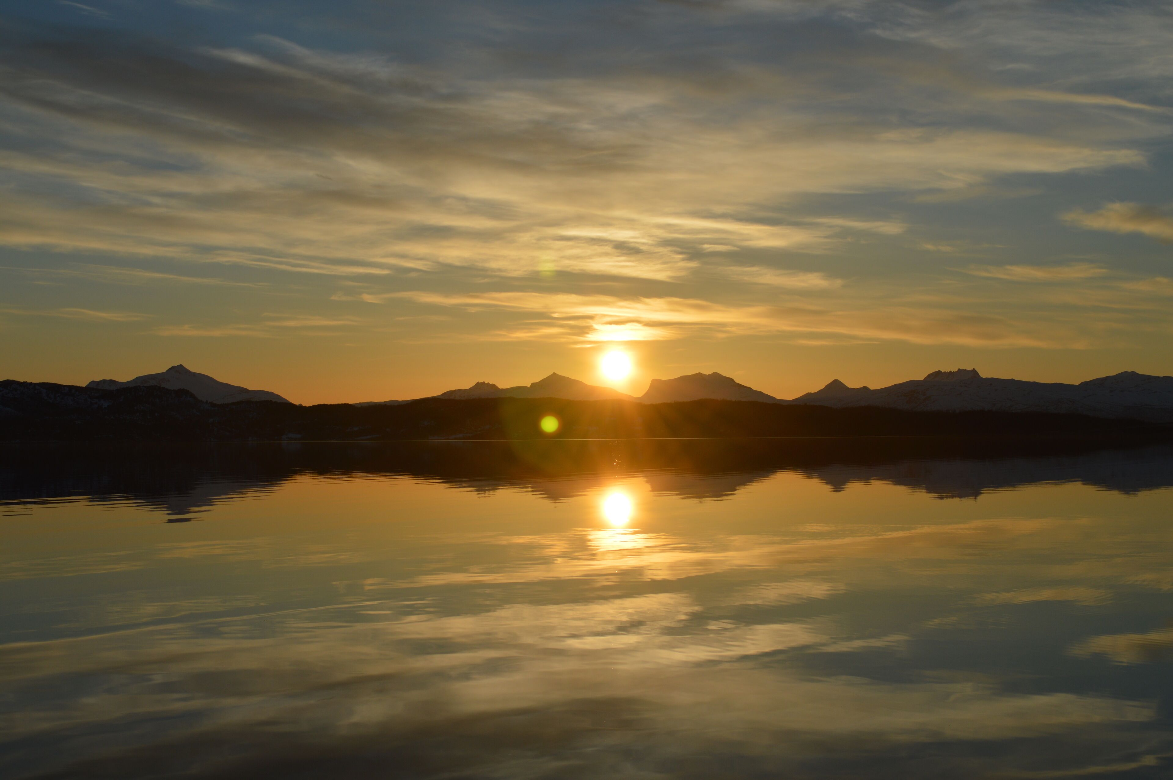En stilla sjö som speglar solen och bergen under solnedgången.