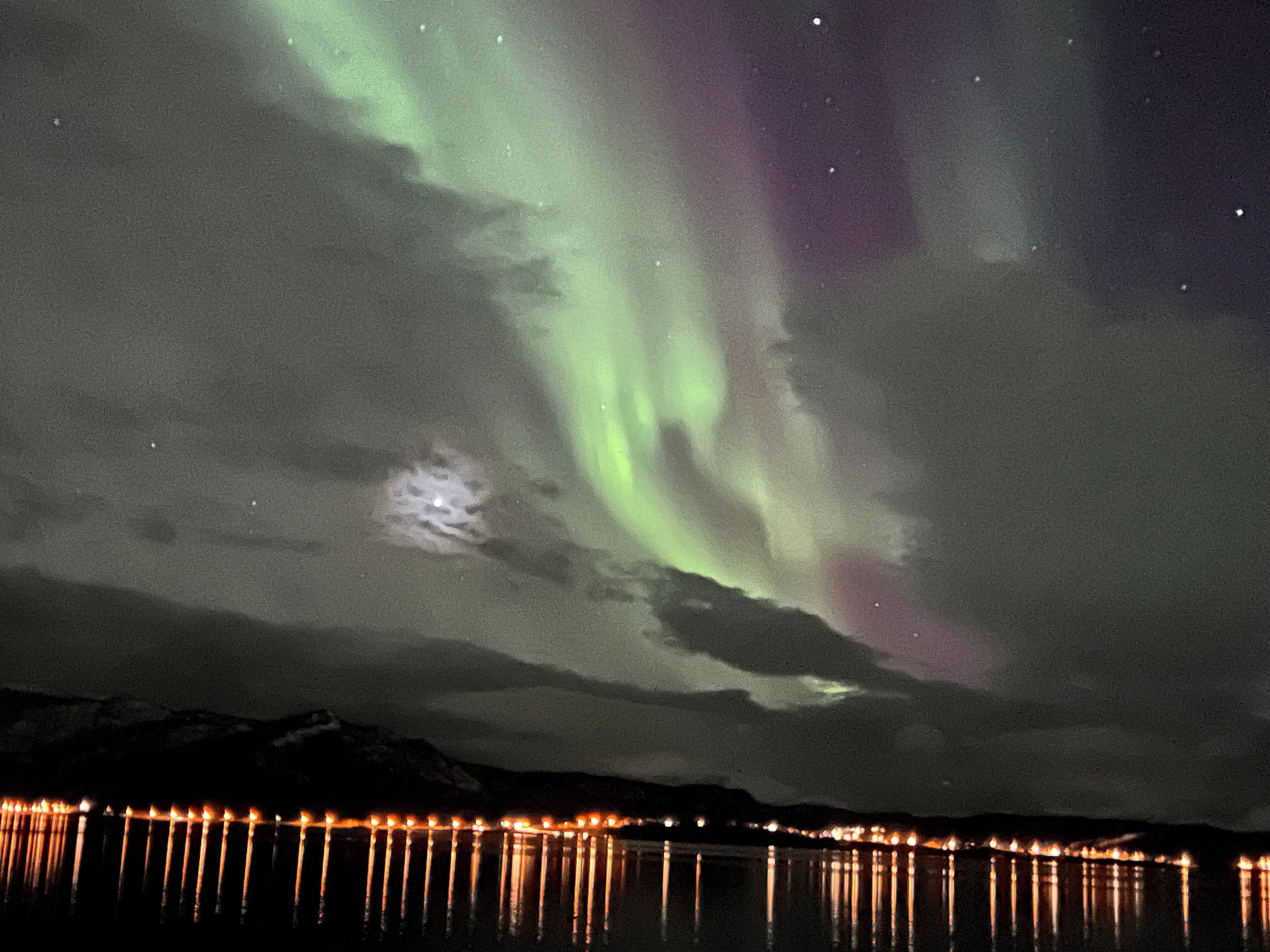 Cielo nocturno con auroras boreales reflejadas en un cuerpo de agua, junto a la luz de la costa distante.