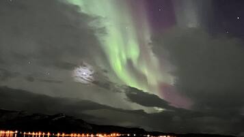 Cielo nocturno con auroras boreales reflejadas en un cuerpo de agua, junto a la luz de la costa distante.