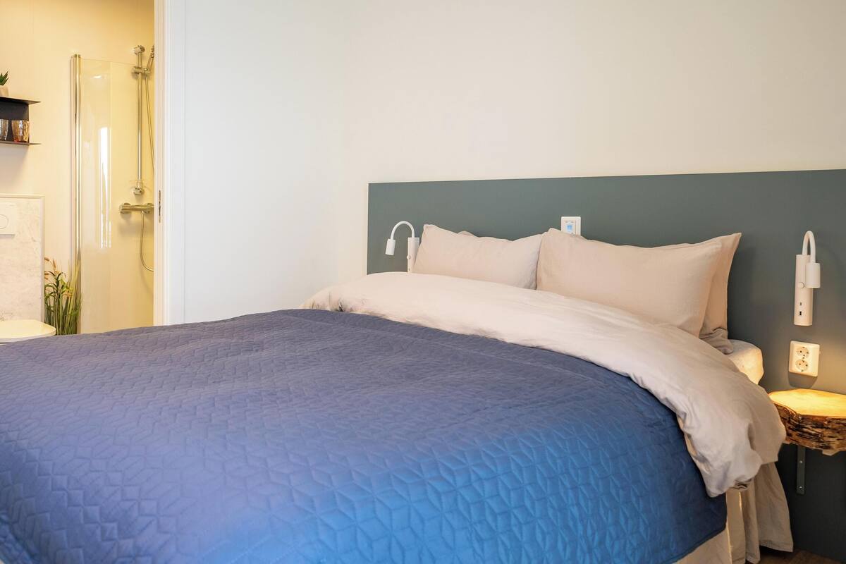 A neatly made bed with a blue quilt, a bedside lamp, and a small wooden stool.