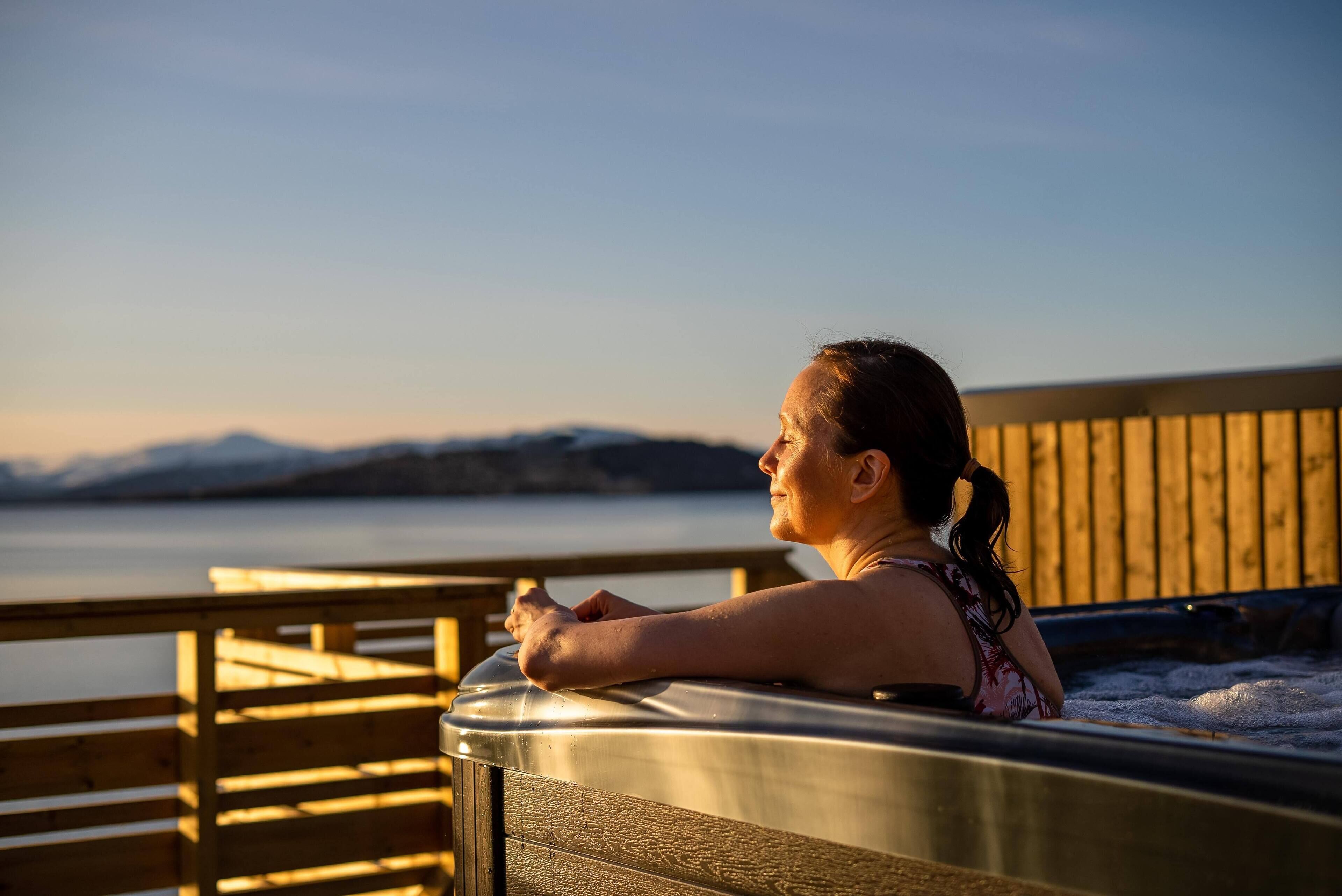 A person relaxing in a hot tub with a wooden deck and a view of a body of water and mountains in the background.