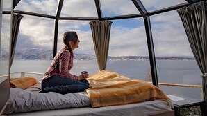 Una habitación con una cama, una persona sentada en ella y un ventanal con vistas a un cuerpo de agua y montañas.