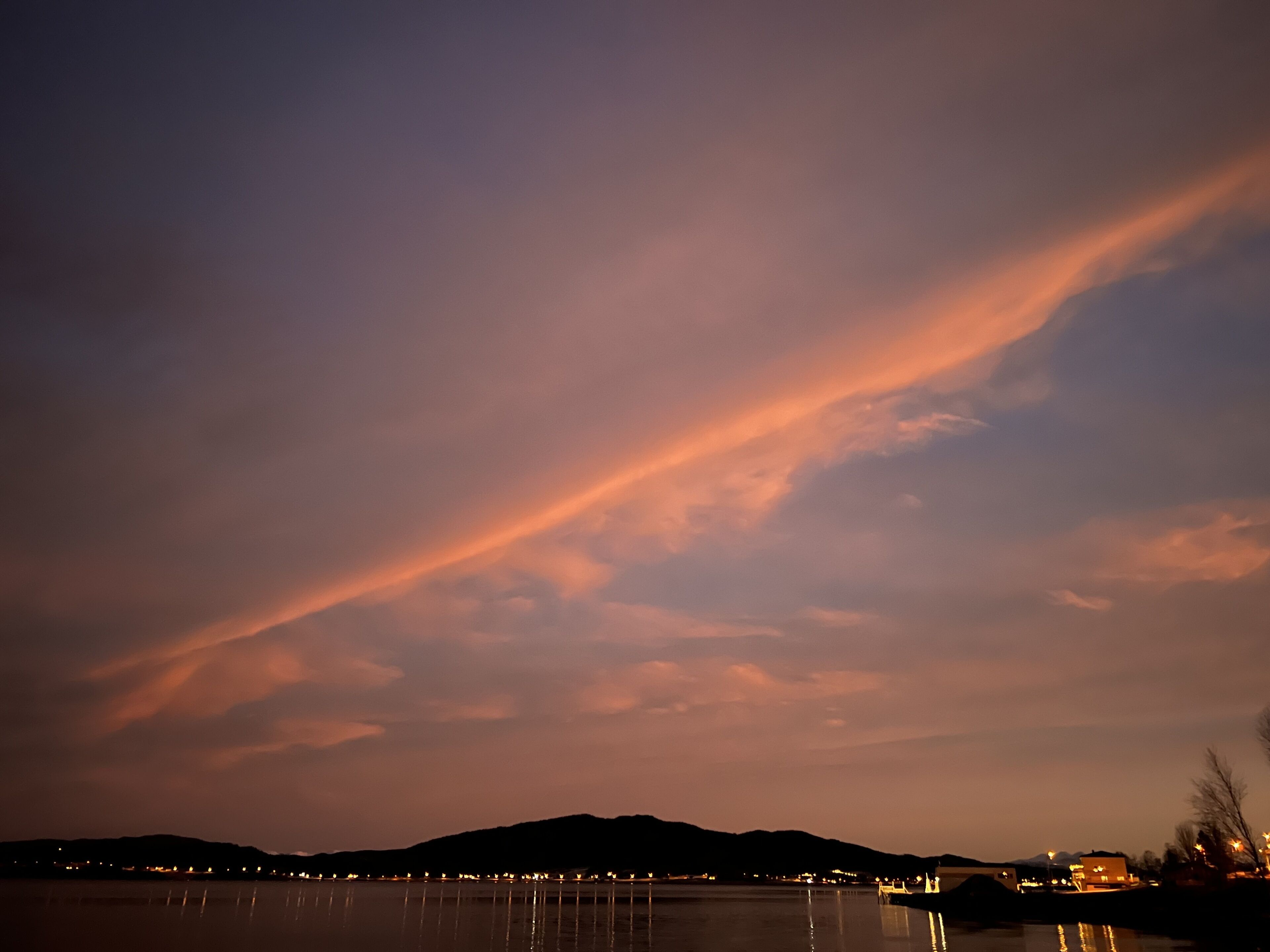 A calm lake at dusk with a mountain in the background and a city skyline on the opposite shore.