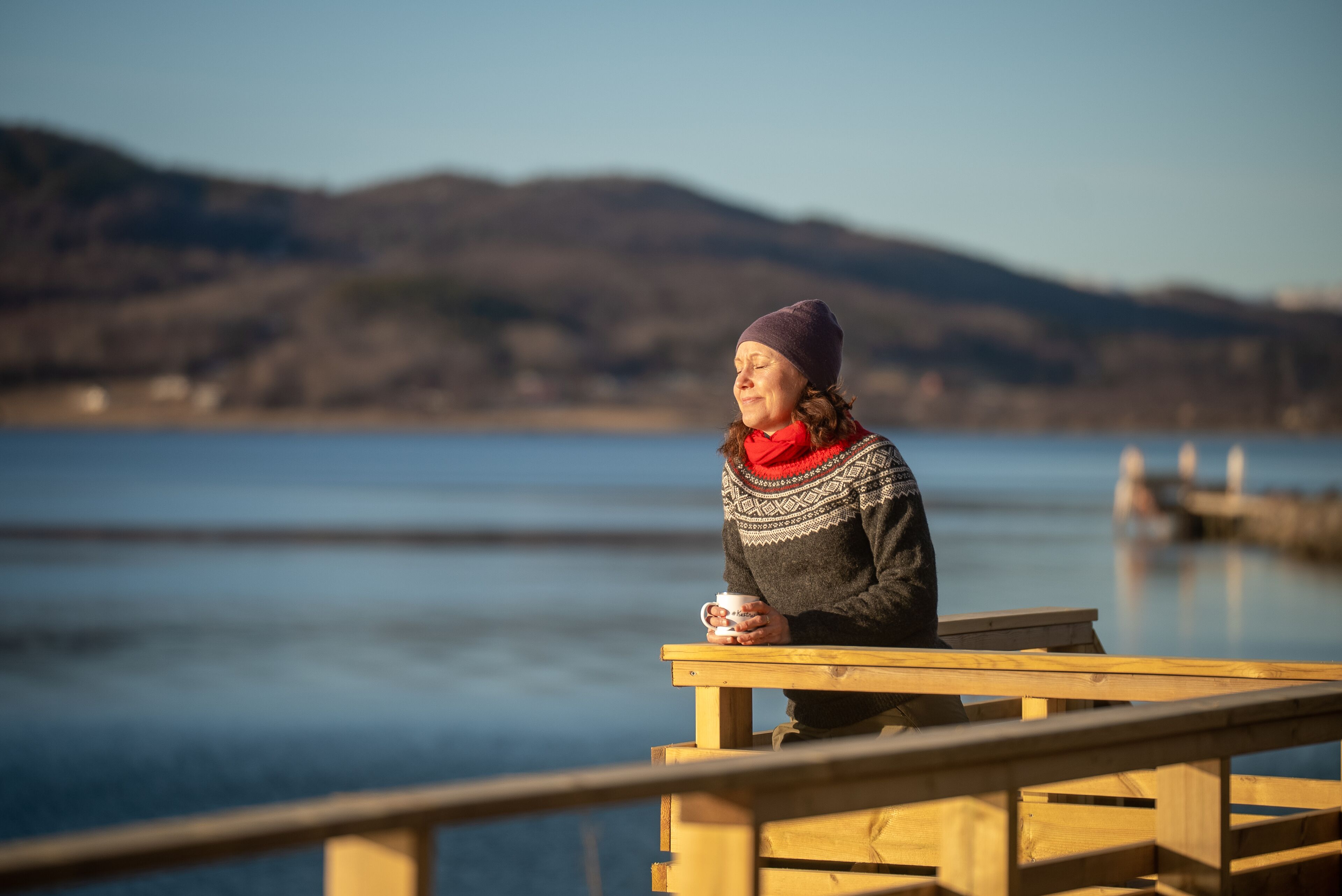 Una persona sentada en un muelle de madera junto a un lago tranquilo con montañas al fondo.