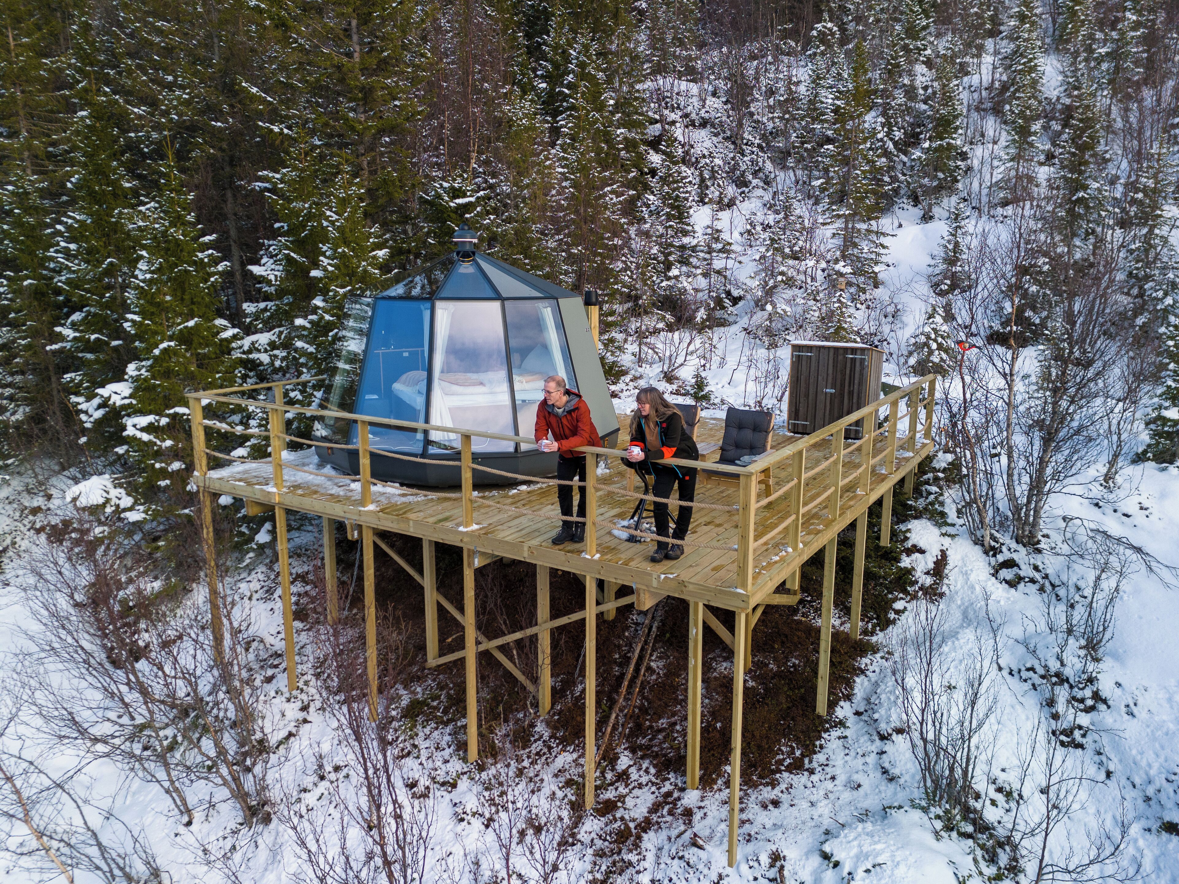 A wooden deck with a glass igloo structure, surrounded by snow-covered trees.