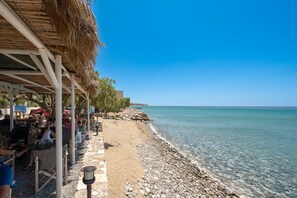 A beachside café with outdoor seating, a thatched roof, and a rocky shoreline.