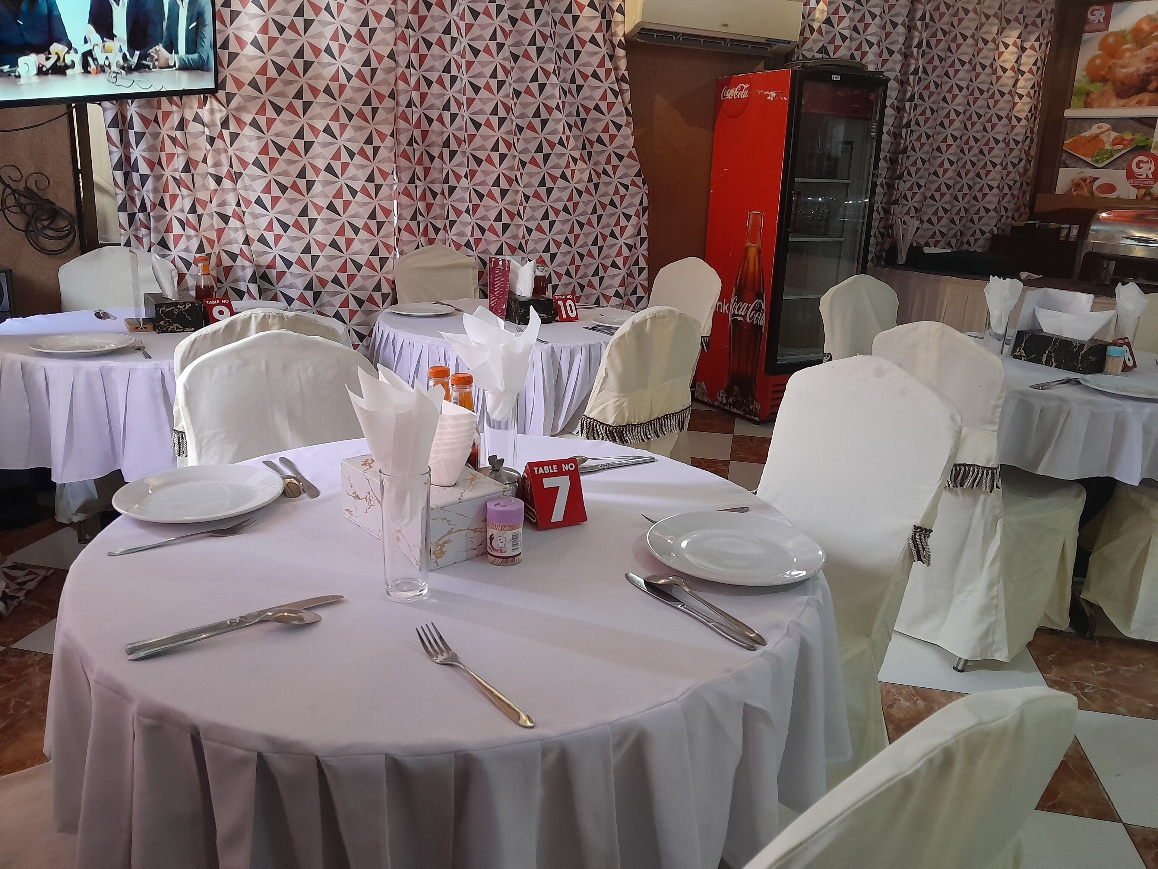 Un área de comedor con mesas redondas dispuestas para cuatro, cada una con manteles blancos, platos, cubiertos y servilletas. Las sillas están cubiertas con tela blanca. Se ve un refrigerador Coca-Cola al fondo.