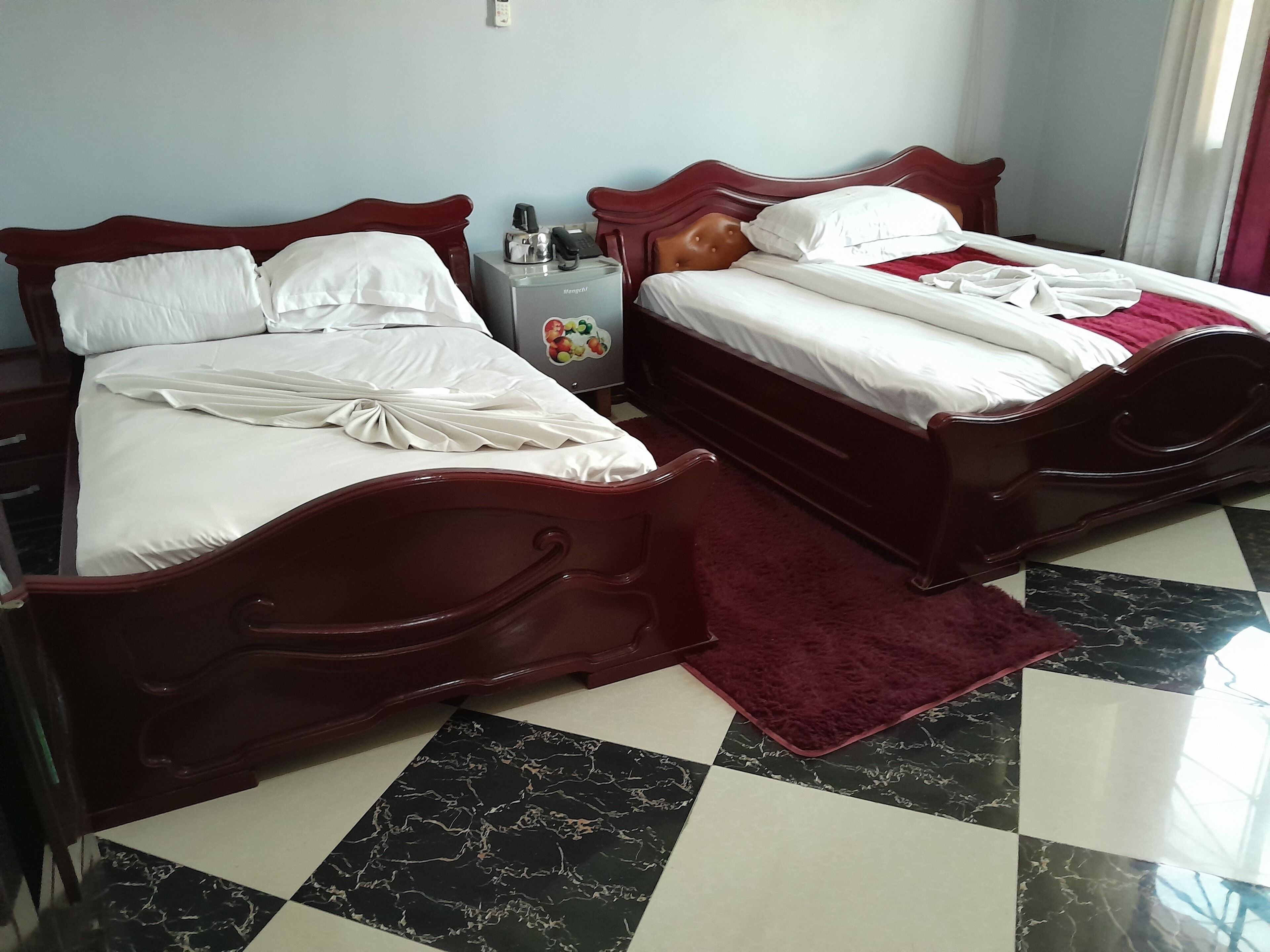 Two wooden beds with white linens, a small table with a coffee maker and phone, and a checkered floor.