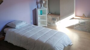Un dormitorio con techo de piedra, una cama con ropa de cama blanca, un estante pequeño con una botella azul y una guitarra colgada del techo.