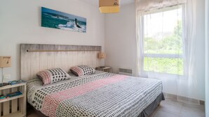 Un dormitorio con cama, mesita de noche, lámpara, ventana con cortinas y un cuadro del océano.
