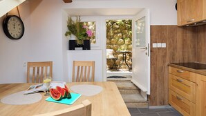A kitchen with wooden cabinets, a dining table with a placemat, chairs, a clock, and a view of the garden through an open door. - Hedera Estate, Hedera A6 (Dubrovnik)