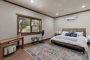 A bedroom with a wooden bed, a bedside table, a nightstand, a window with blinds, a standing fan, and a wall-mounted air conditioner. - Ranch Retreat Creek View - Pool Table and Fire Pit (Fredericksburg)