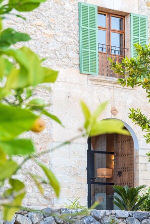 A stone building with a wooden door and green shutters, a balcony with a railing, and a stone wall. - Agroturisme Subies (Selva)