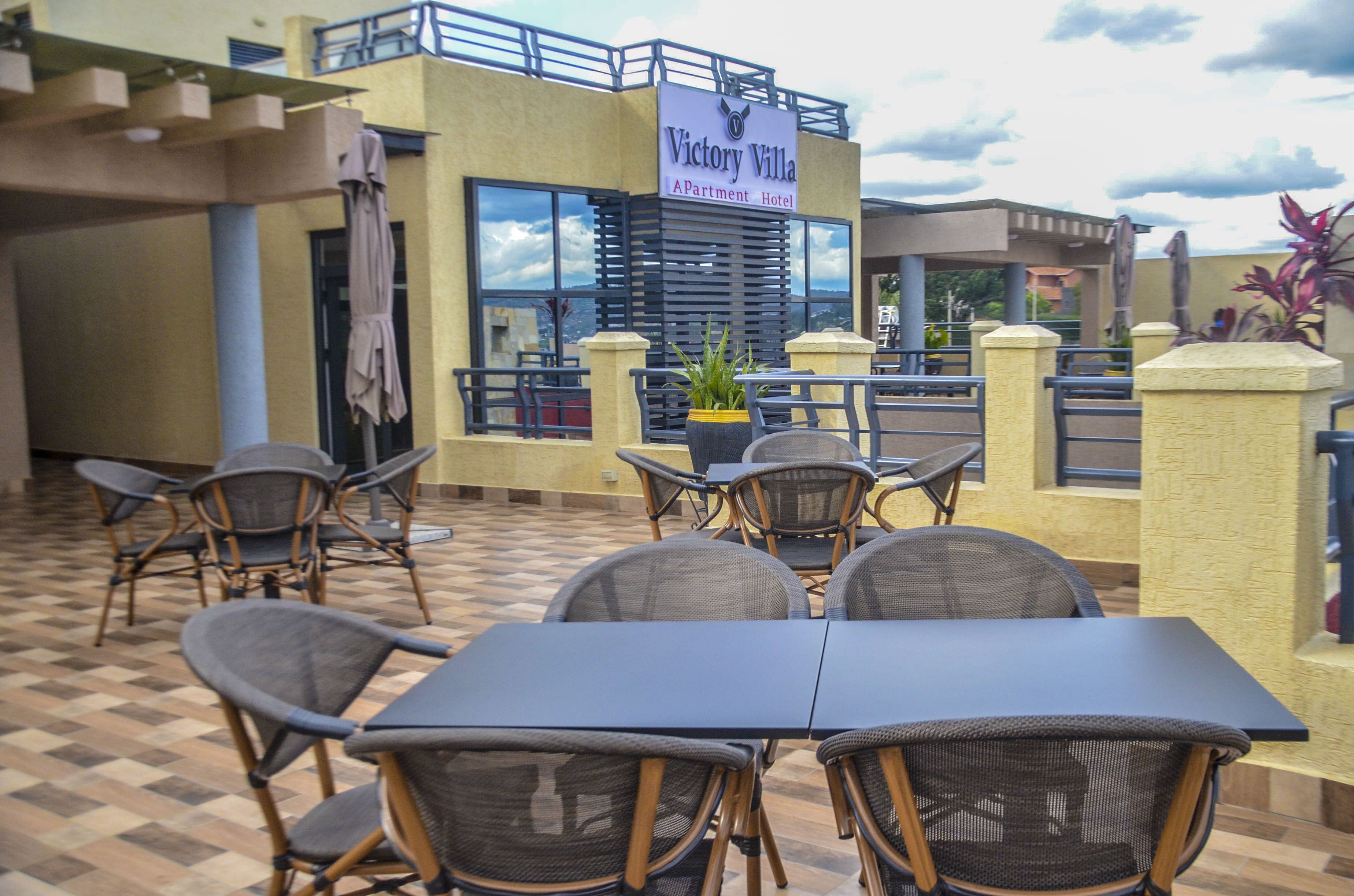 An outdoor patio area with tables and chairs, a sign for Victory Villa Apartment Hotel, and a view of the surrounding landscape.