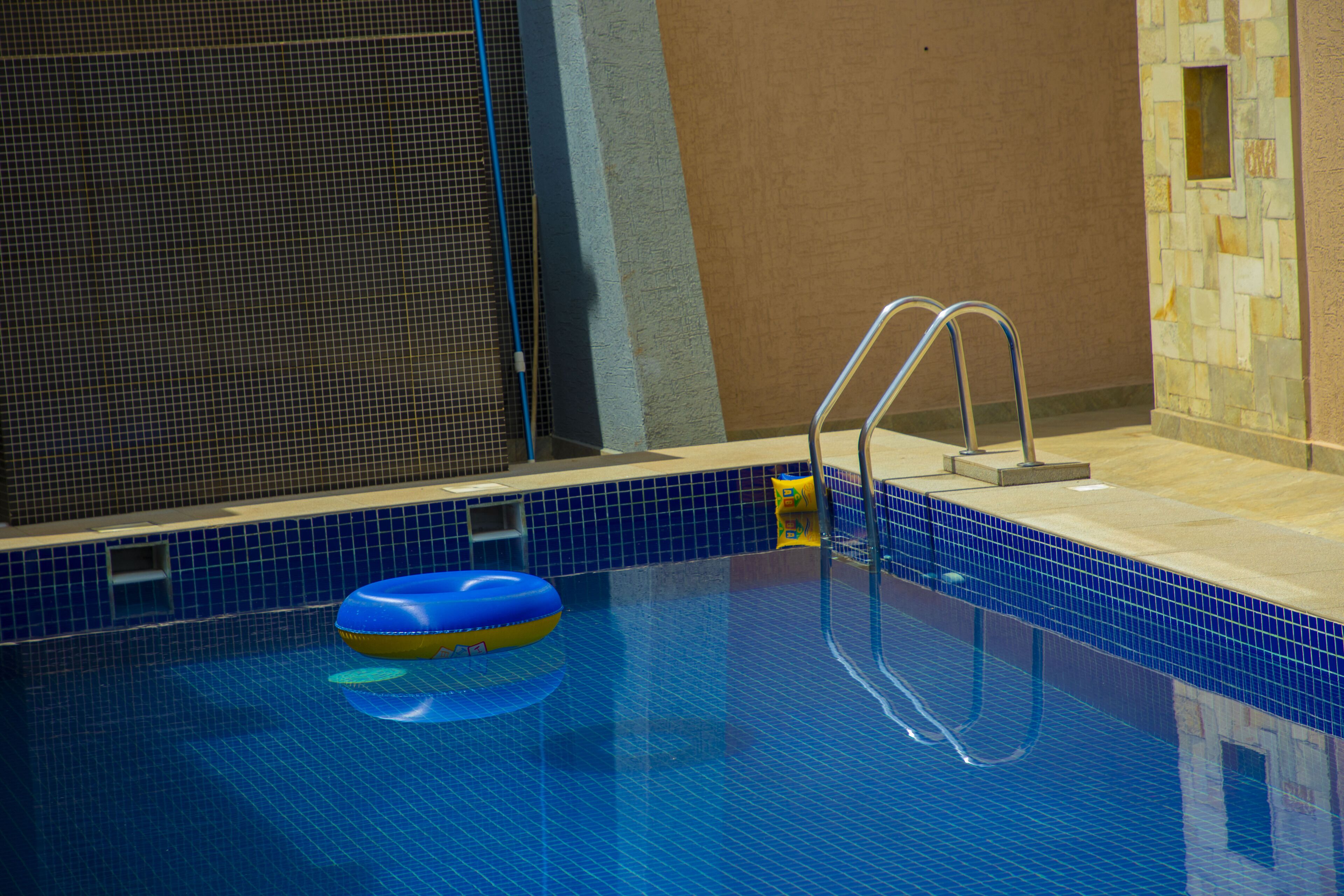 An indoor swimming pool with a blue tiled floor, a pool ladder, and a blue and yellow floating ring.