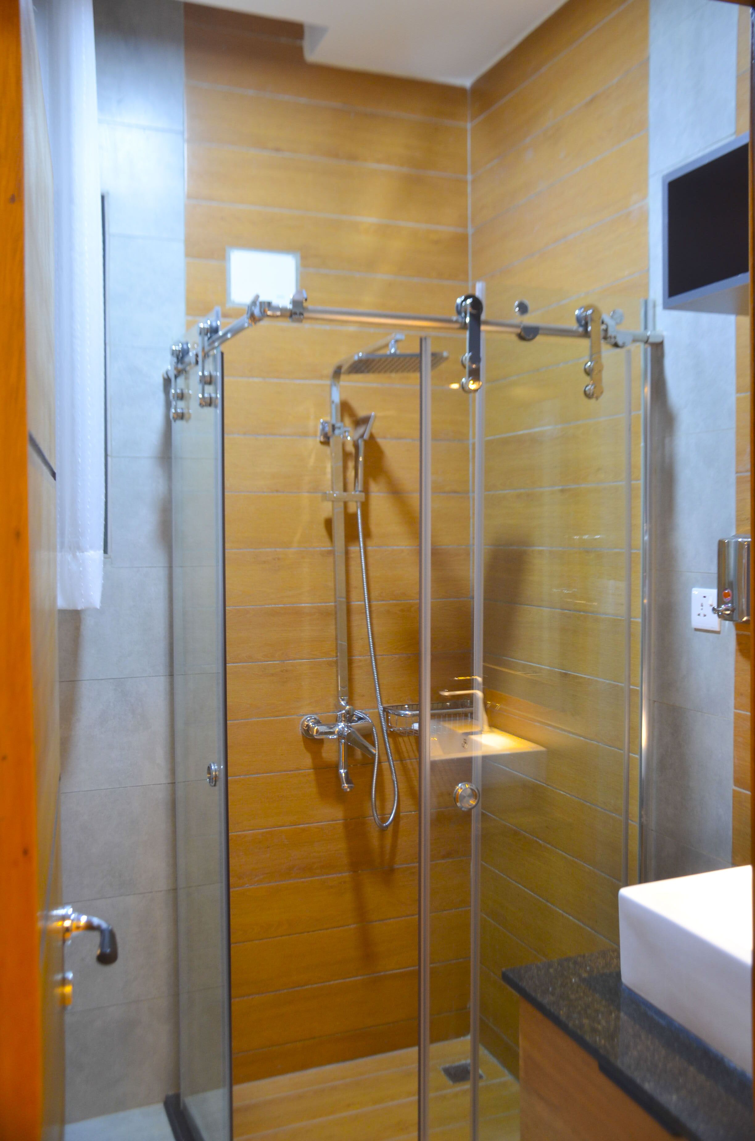 A glass-enclosed shower with a wooden panel wall, a sink, and a flat-screen TV mounted on the wall.