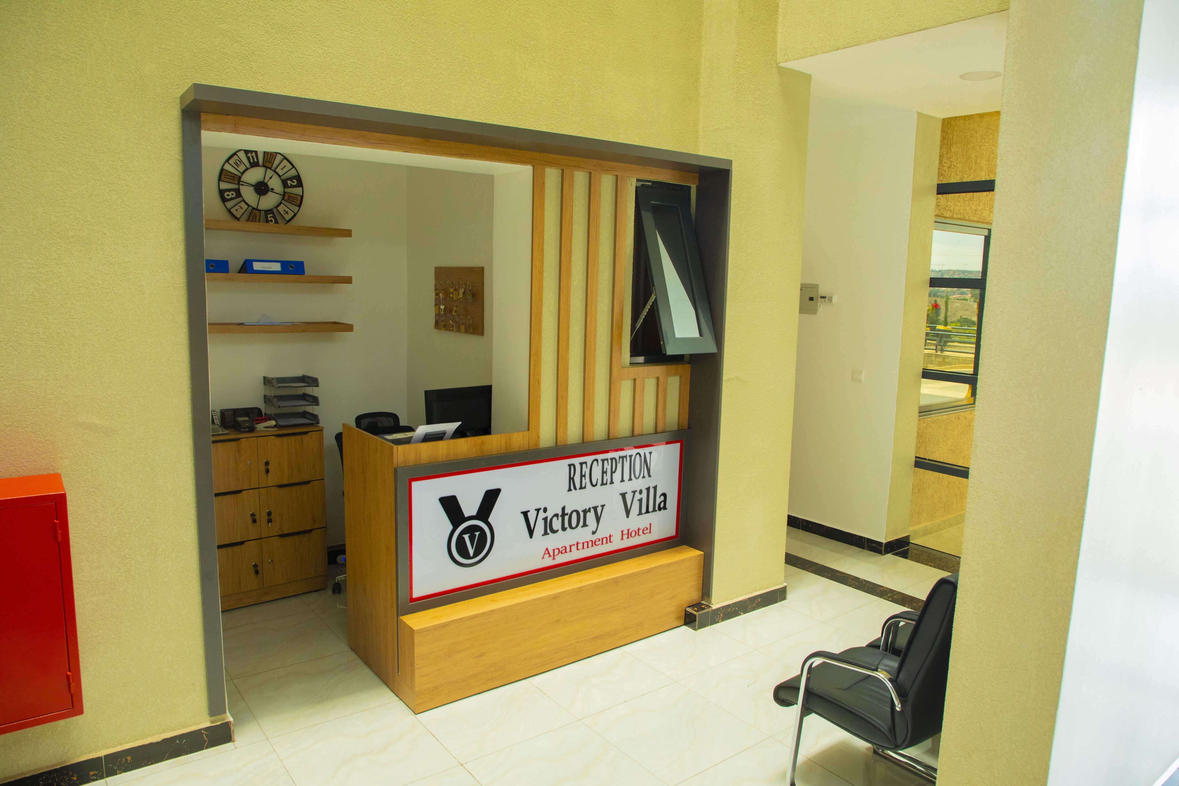 A reception desk with a sign for Victory Villa Apartment Hotel, wooden shelving, a clock, and a black chair.