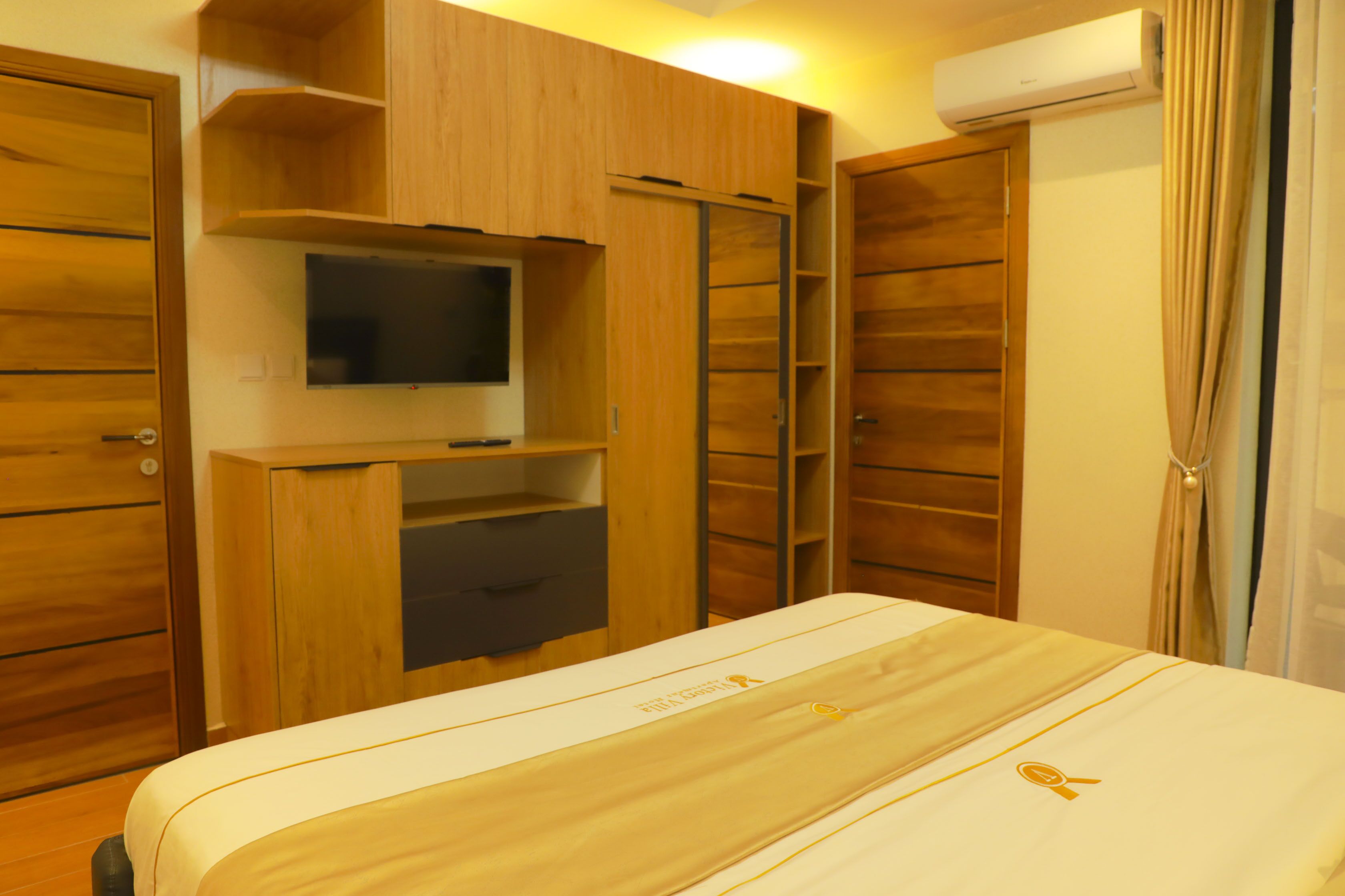 A hotel room with a bed, built-in TV, wooden wardrobe, and a sliding door closet.
